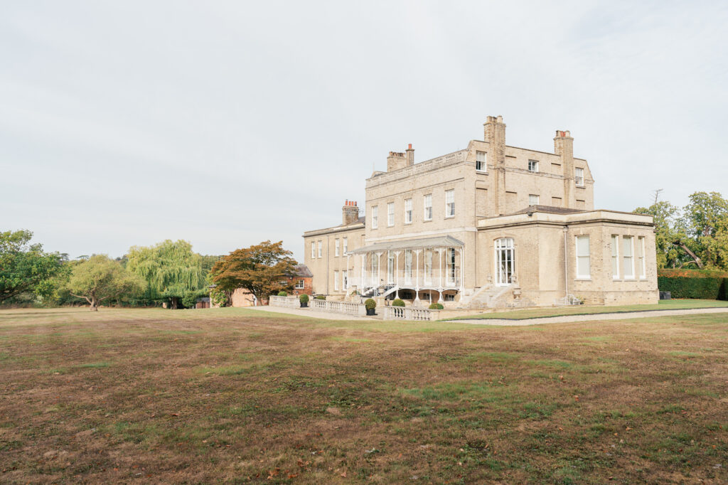 A large, elegant, light-colored mansion with tall windows sits on a spacious, grassy lawn surrounded by trees under a bright, partly cloudy sky.