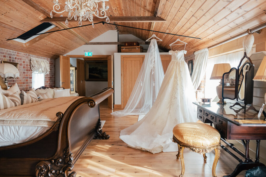 A bridal suite with wooden walls and ceiling, a vintage bed, and a vanity table. A wedding dress and long veil hang near double doors, illuminated by natural light from large windows.