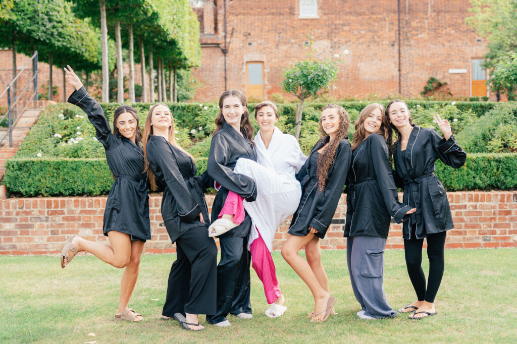 Seven women pose happily outdoors in front of a garden and brick building. Six wear matching black robes, and one in the center wears a white robe with pink pants, lifting her leg playfully. They all smile at the camera.