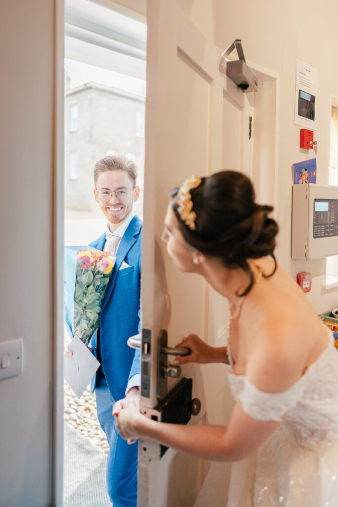 A bride in a white dress opens a door while smiling at a groom in a blue suit holding a bouquet of flowers and an envelope, standing outside and smiling warmly at her.