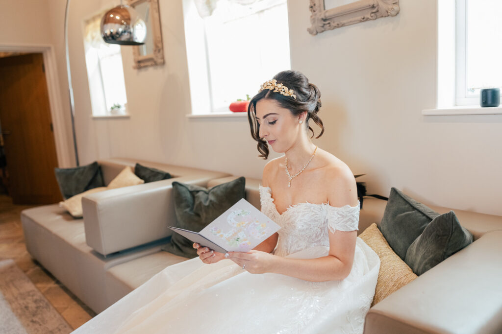 A bride in an off-the-shoulder white gown and jeweled headband sits on a sofa, reading a card. The room is bright with natural light from the windows and decorated with light-colored cushions and mirrors.