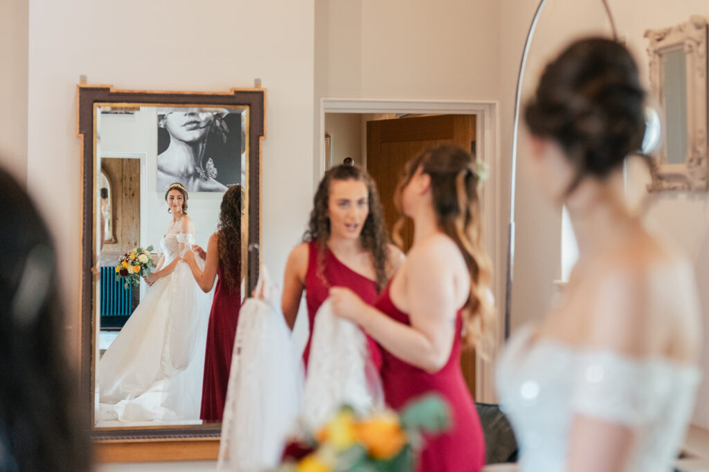 A bride in a white dress is seen in a mirror holding a bouquet, while two bridesmaids in red dresses stand nearby, talking and holding a garment in a softly-lit room.
