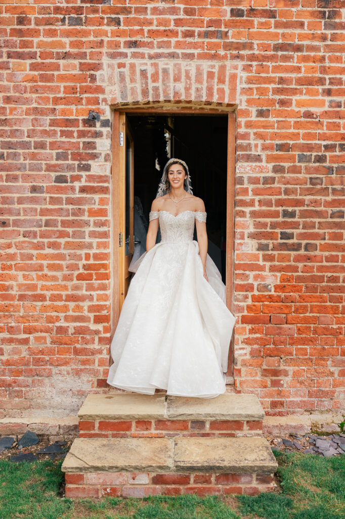 A bride in an off-the-shoulder white wedding dress stands smiling in a doorway set in a red brick wall, with stone steps and green grass in front.