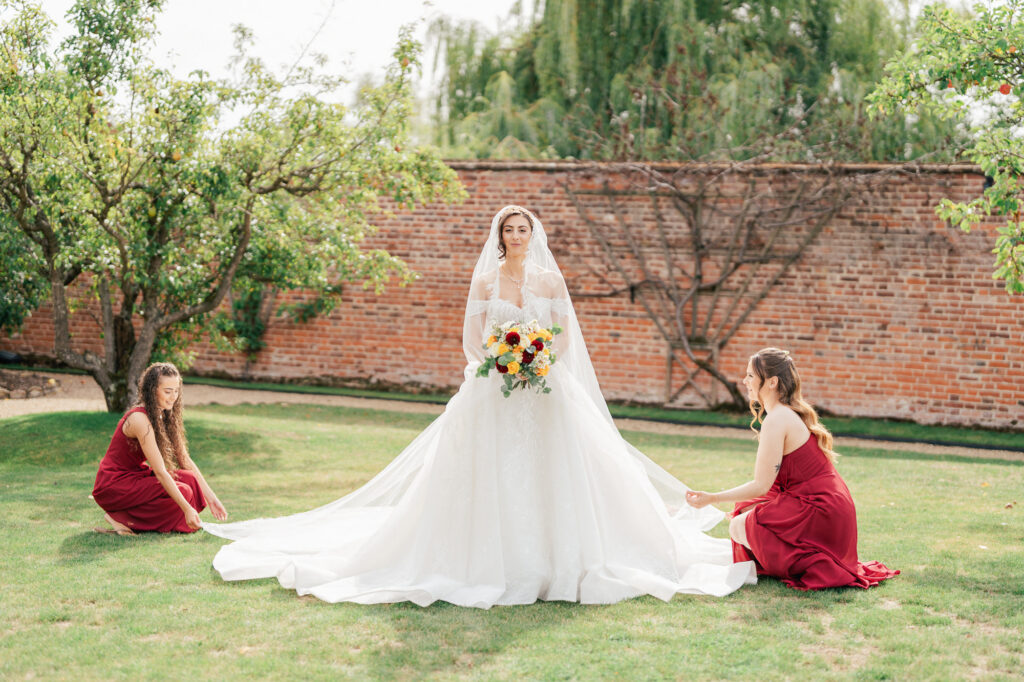 A bride in a white gown and veil holds a colorful bouquet while standing on grass. Two bridesmaids in red dresses kneel beside her, arranging her dress’s train. There is a brick wall and green trees in the background.