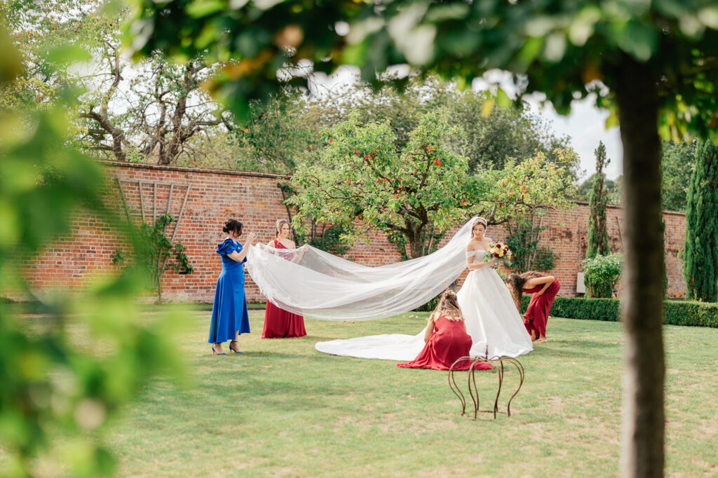A bride in a white gown stands on a lawn as bridesmaids in red dresses and a woman in blue help arrange her long veil. They are outdoors in a garden with green trees and a brick wall in the background.