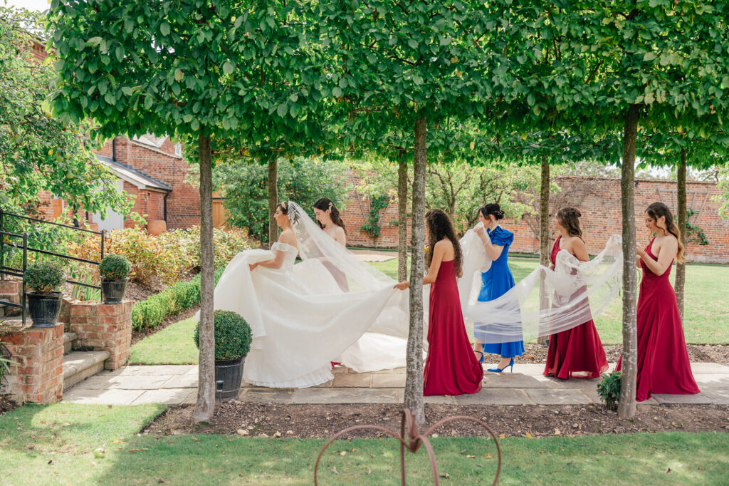 A bride in a white gown stands under leafy trees as bridesmaids in burgundy dresses and a woman in blue arrange her long train and veil in a sunny garden with brick walls in the background.