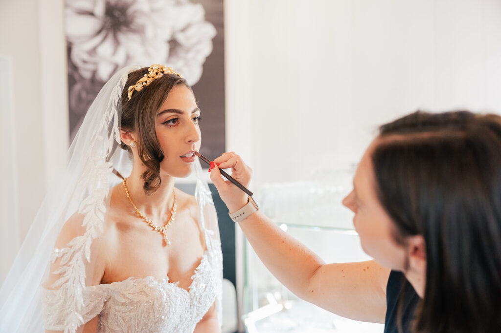 A bride in a white wedding dress and veil has her makeup done by an artist applying lipstick. The bride sits calmly, with a floral wall art in the background.