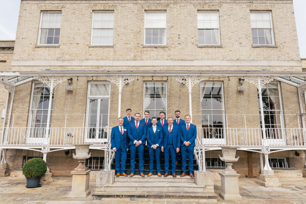 Seven men in matching blue suits and red ties stand on stone steps in front of a large, elegant building with tall windows and white iron railings, posing for a group photo.