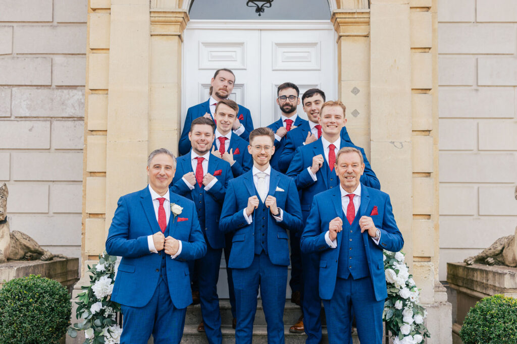 A group of eight men in matching blue suits with red ties pose smiling on stone steps in front of a grand building. The groom stands in the center, holding his lapels, surrounded by his groomsmen.