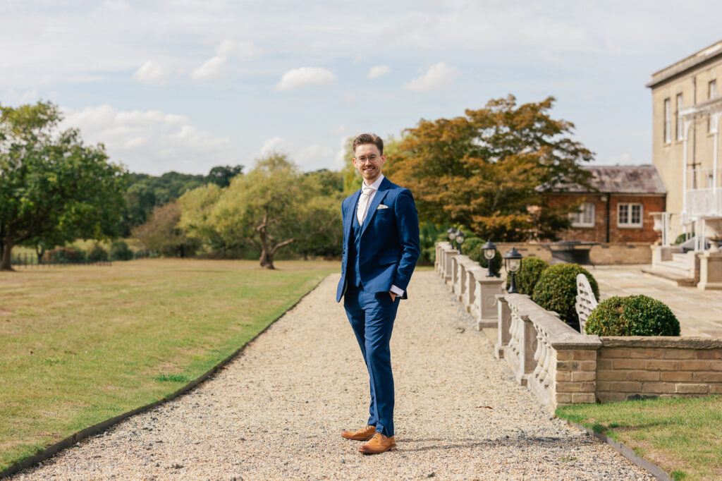 A man in a blue suit stands smiling on a gravel pathway in a garden with trees, grass, and a large building in the background on a sunny day.