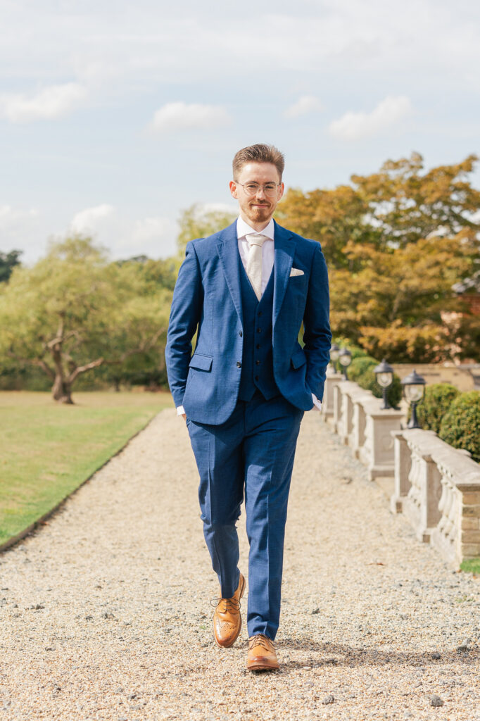 A man in a blue suit, white shirt, and brown shoes walks confidently outside on a gravel path, with greenery and trees in the background under a partly cloudy sky.