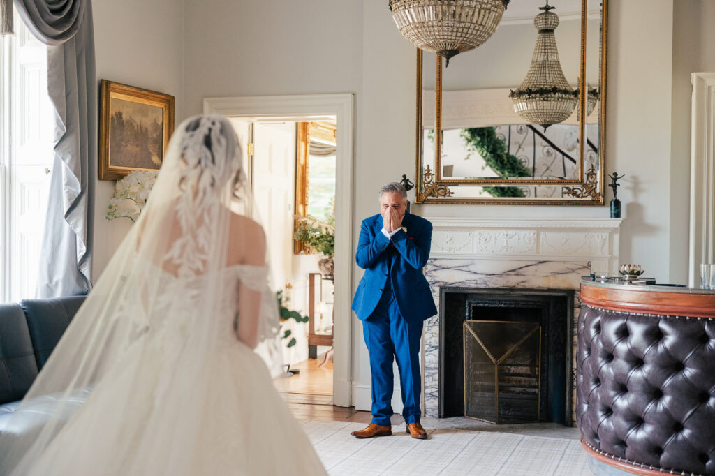 A bride in a white gown faces an older man in a blue suit who looks surprised and emotional, standing in a bright, elegant room with a chandelier and large mirror.