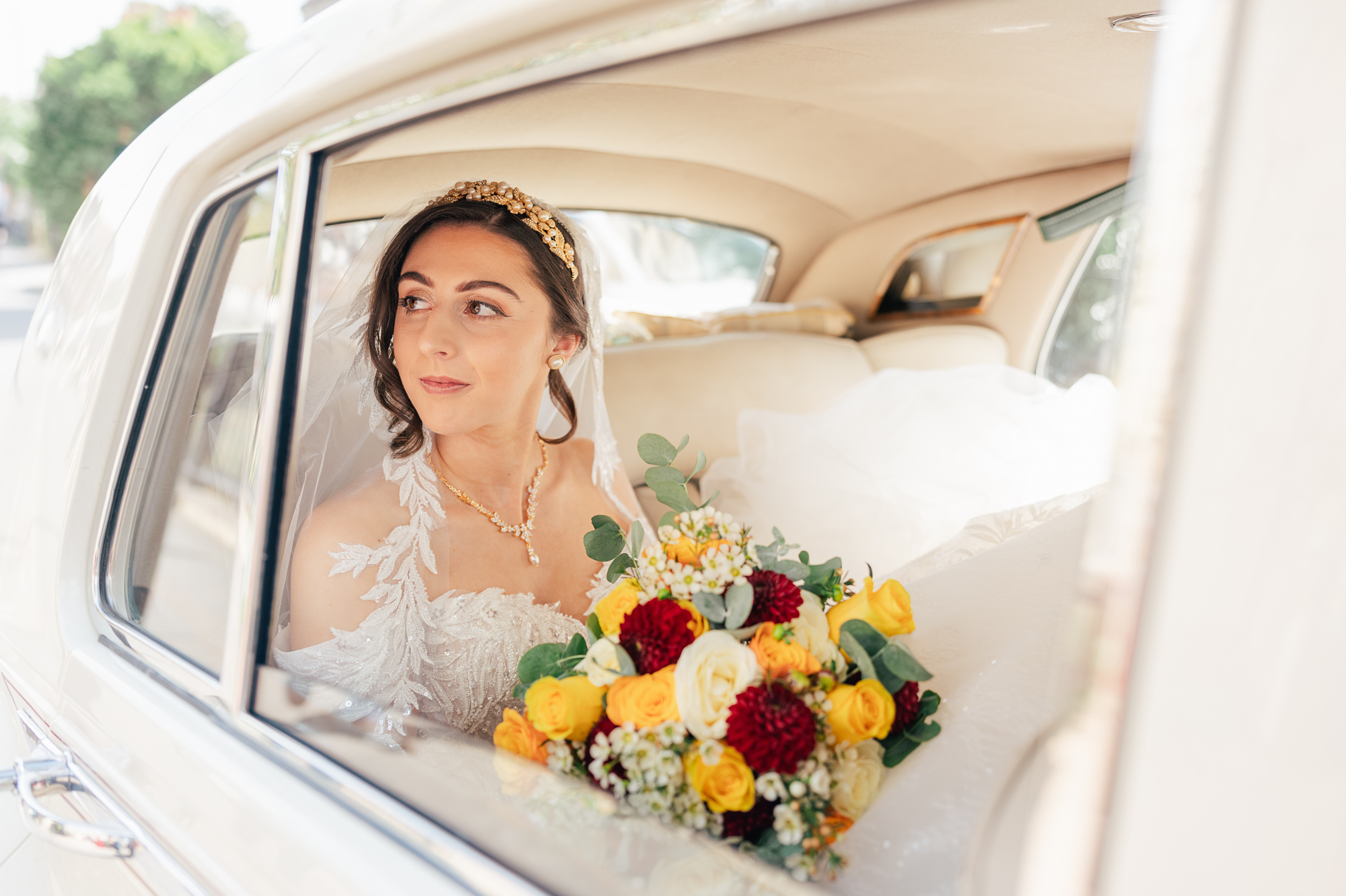 A bride in a white dress holds a bouquet of red, yellow, and white flowers while sitting in the back seat of a vintage white car, looking thoughtfully out the window.