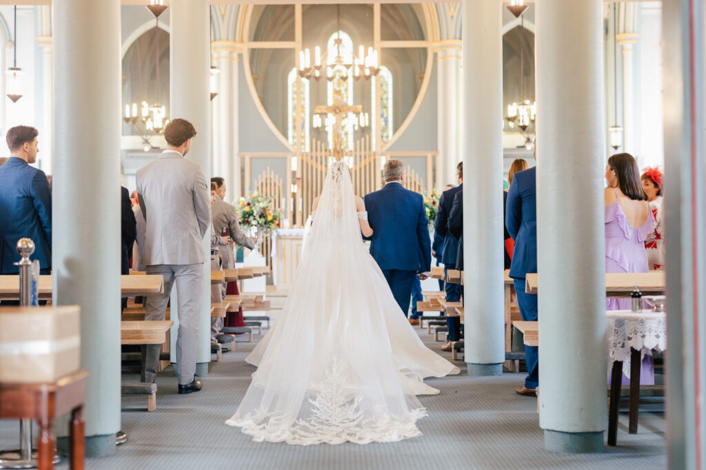 Bride in a long white gown and veil walks down the aisle with an older man in a blue suit inside a bright, elegant church, as guests stand and watch from both sides.