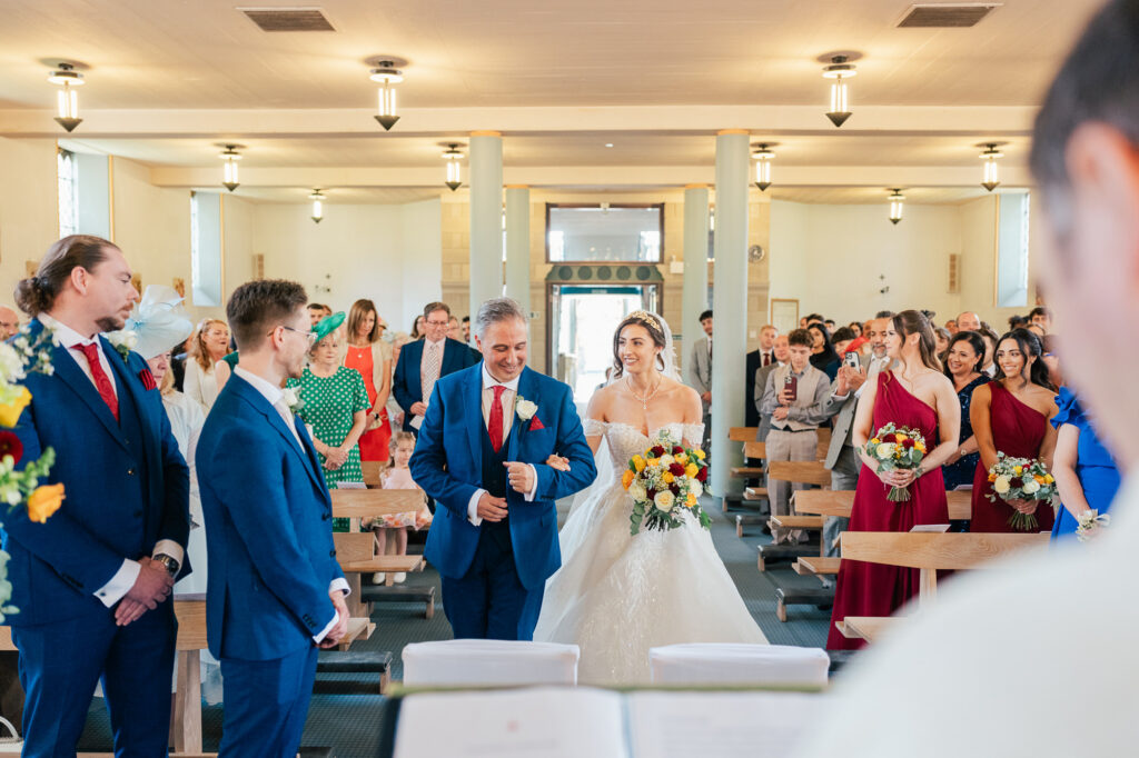 A smiling bride in a white gown walks down the aisle with her father in a bright, modern church filled with seated guests. Bridesmaids in red dresses and groomsmen in blue suits watch joyfully.