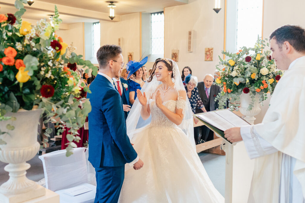 A bride and groom stand facing each other, smiling joyfully during their wedding ceremony in a bright church, as a priest officiates and guests watch from the pews. Large flower arrangements decorate the scene.