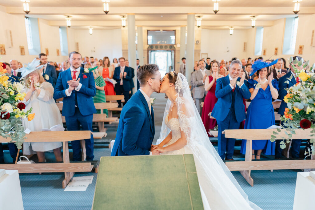 A bride and groom kiss at the altar in a brightly lit church, while guests in colorful attire stand, clap, and smile in celebration behind them.