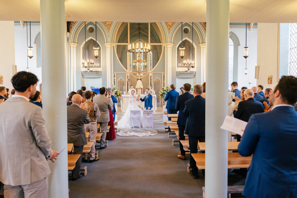 A bride and groom stand at the altar in a church, surrounded by wedding guests seated in pews. The church features tall columns, arched architecture, and large chandeliers, creating an elegant atmosphere.