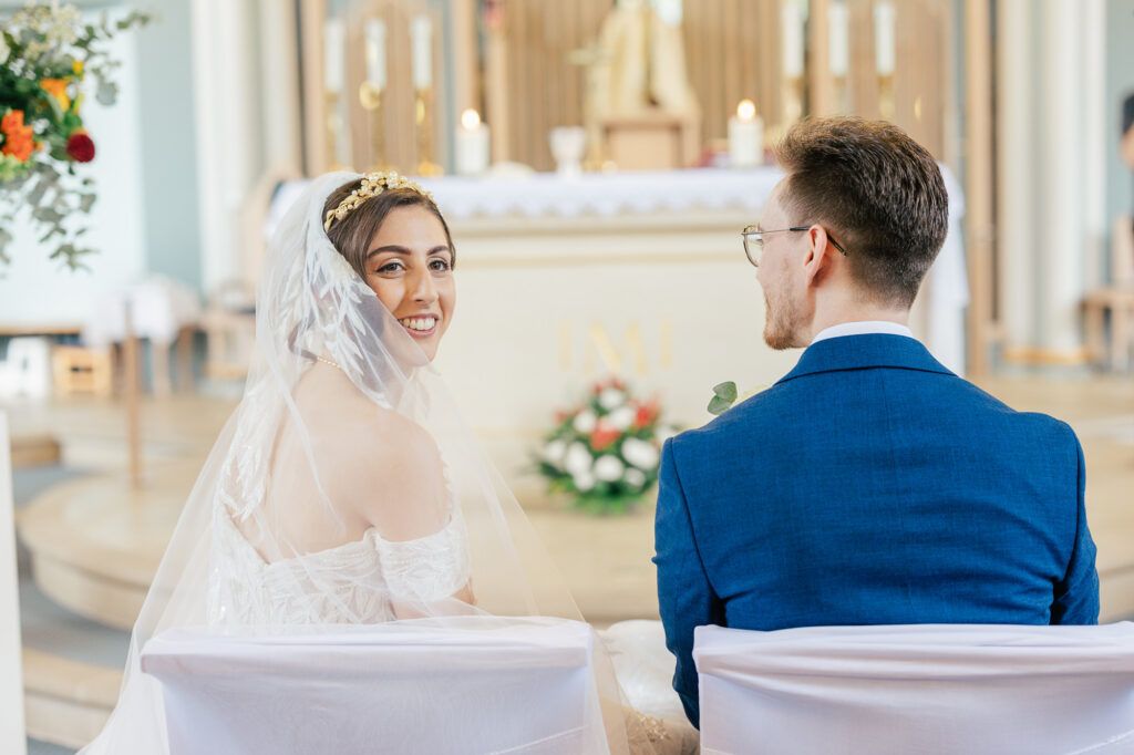 A bride in a white dress and veil smiles at the camera while sitting beside the groom, who wears a blue suit, in a church decorated with flowers and candles.