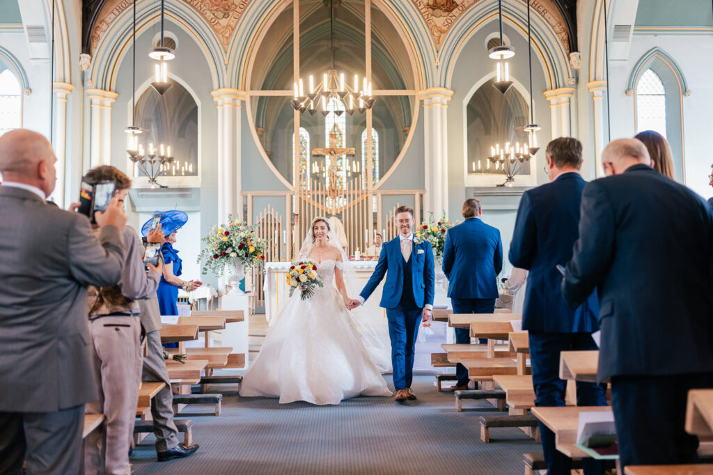 A bride and groom walk hand-in-hand down the aisle of a church after their wedding ceremony, smiling as guests in formal attire stand, watch, and take photos. The church has high arched ceilings and chandeliers.