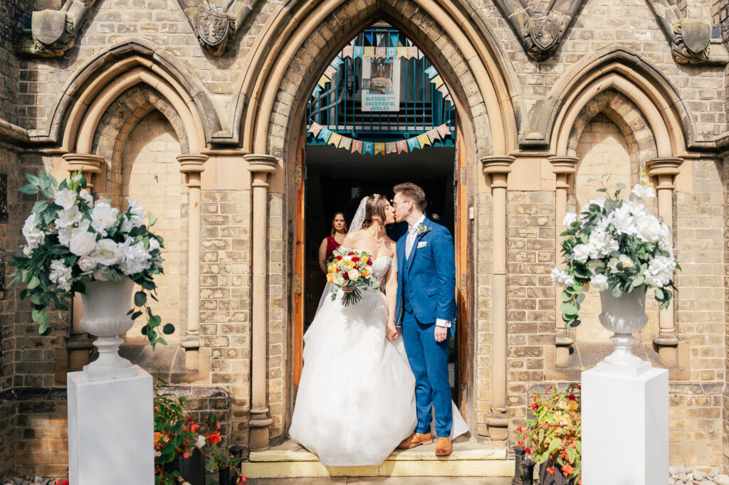 A bride and groom stand in the doorway of a brick church, sharing a kiss. The bride holds a bouquet and wears a white gown; the groom is in a blue suit. Floral arrangements and a bridesmaid appear in the background.