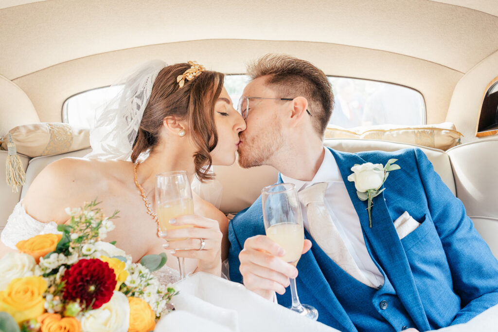 A bride and groom sit in the back of a car, dressed in wedding attire, kissing while holding champagne glasses. The bride holds a bouquet of flowers and both appear joyful and celebratory.