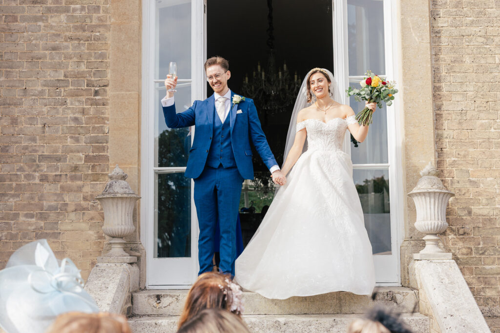 A newlywed couple stands on stone steps outside a building. The groom, in a blue suit, raises a glass and smiles, while the bride, in a white gown, holds a bouquet and their hands. Guests watch from below.