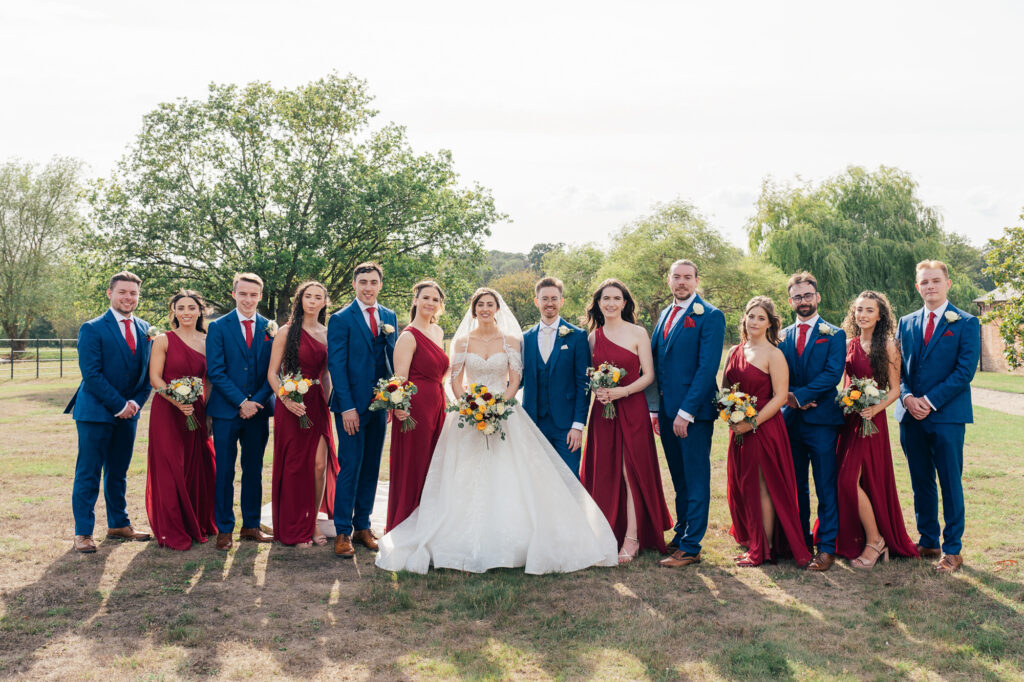 A wedding party poses outdoors. The bride and groom stand in the center, surrounded by bridesmaids in burgundy dresses and groomsmen in blue suits, all holding bouquets, with trees and grass in the background.