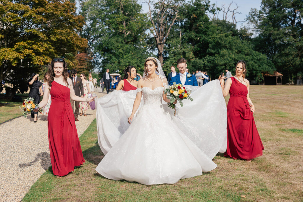 A bride in a white gown is smiling as she walks outdoors, surrounded by bridesmaids in red dresses and a man in a blue suit. The bridesmaids hold her dress and bouquets. The group is walking on grass near a gravel path with trees in the background.