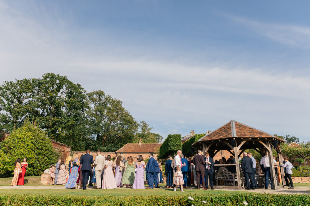 A group of people dressed in formal attire gather outdoors near a wooden pavilion on a sunny day. The scene is surrounded by greenery, trees, and a clear blue sky.