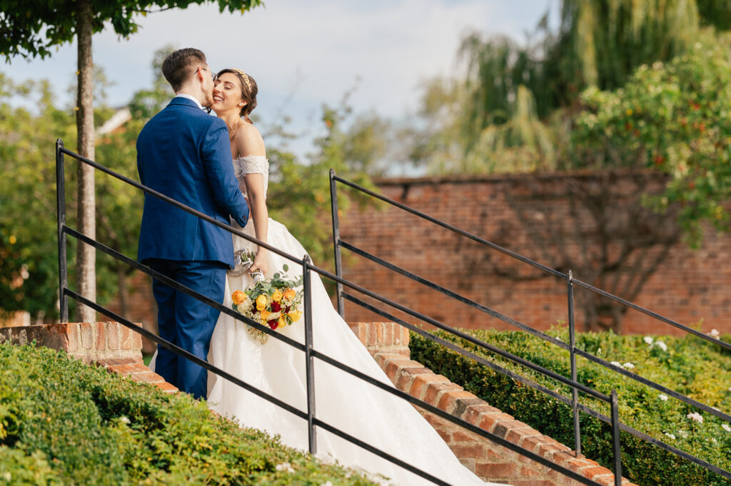A bride in a white gown holding a bouquet smiles at a groom in a blue suit as they stand together on an outdoor brick staircase, surrounded by greenery and trees under a blue sky.