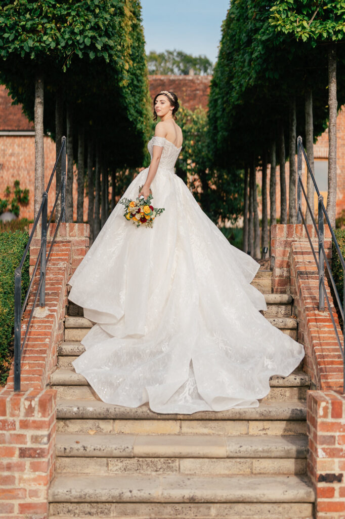 A bride in a flowing white gown stands on outdoor stone steps, holding a bouquet of yellow and orange flowers, with green trees and brick walls in the background.