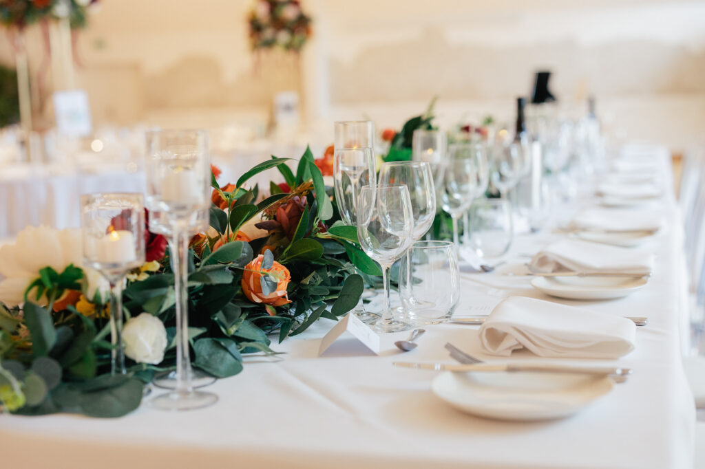 Elegant table setting with white linens, glassware, plates, and silverware. Greenery and autumn-colored flower centerpiece runs along the table. Candles in glass holders add a warm, festive touch.