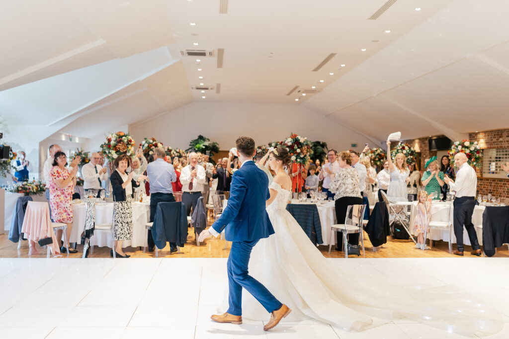 A bride and groom walk hand-in-hand into a bright, elegant wedding reception as guests stand and applaud. The room is decorated with large floral arrangements and round tables.