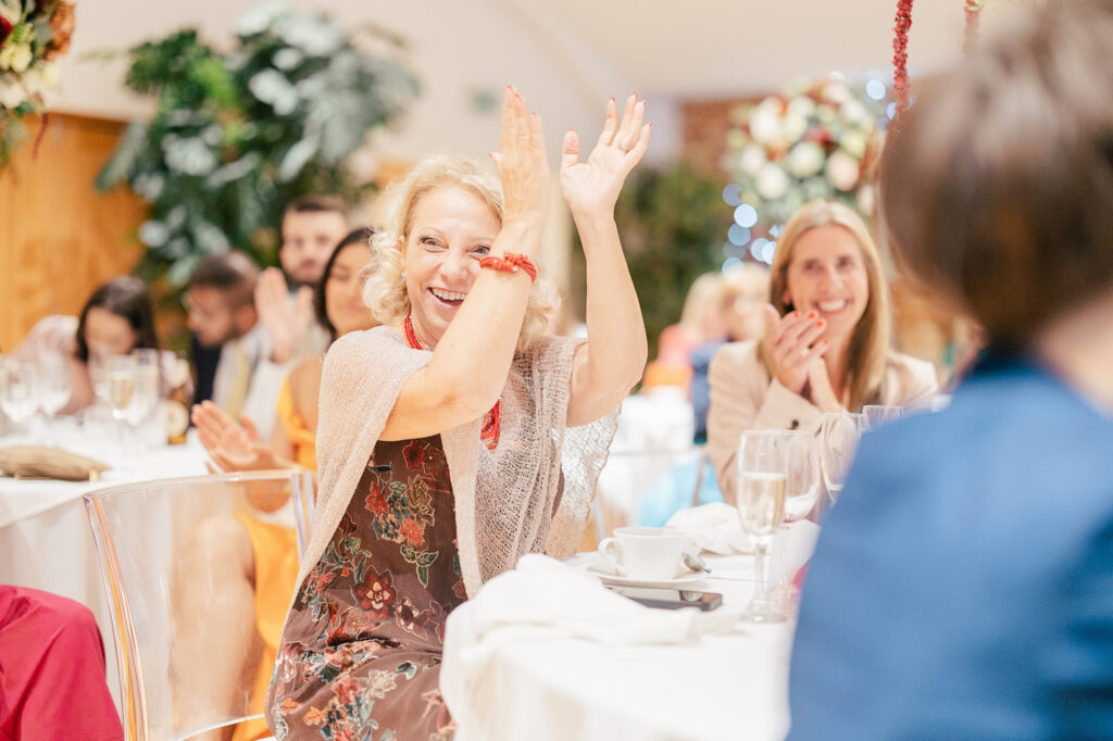 A woman in a floral dress smiles and claps enthusiastically at a formal event, seated at a table with white linens alongside other applauding guests in a bright, festive setting.