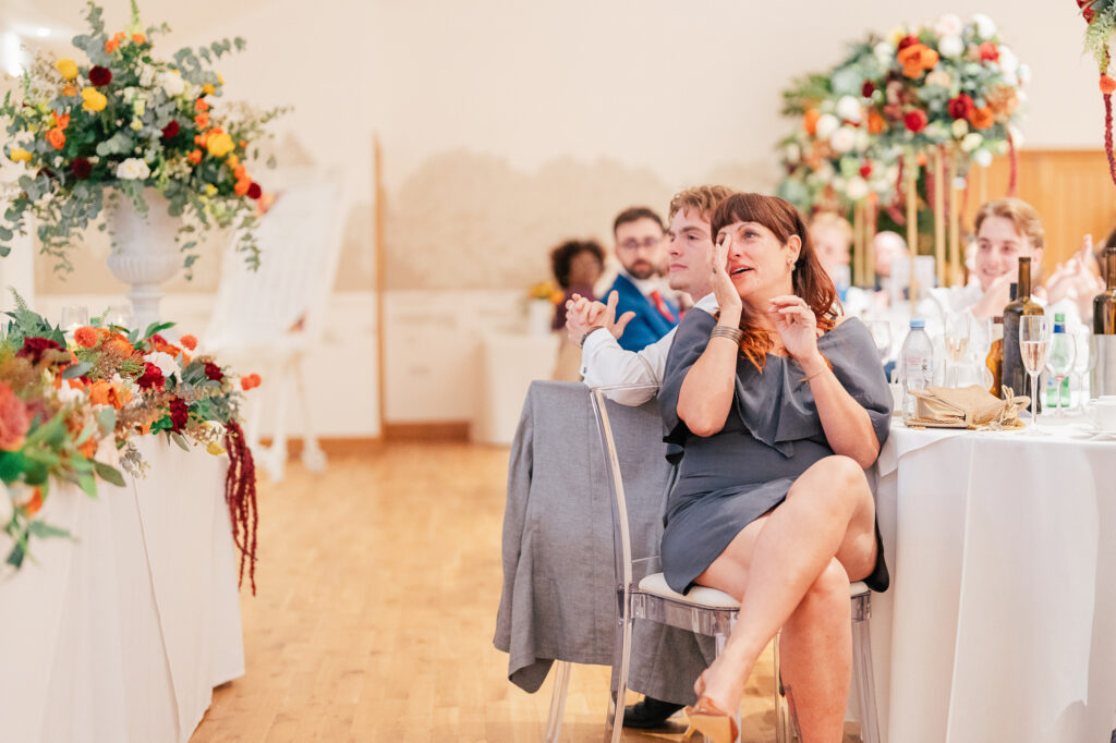 A woman in a gray dress sits at a round table decorated with flowers, smiling and clapping during a formal event. Guests around her watch attentively, and the room is filled with bright floral arrangements.