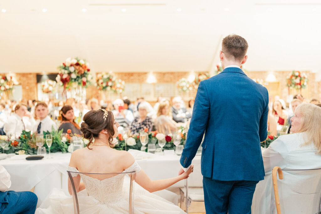 A bride and groom, seen from behind, hold hands while standing at the head table during a wedding reception. Guests sit around decorated tables with floral centerpieces and glasses of champagne.