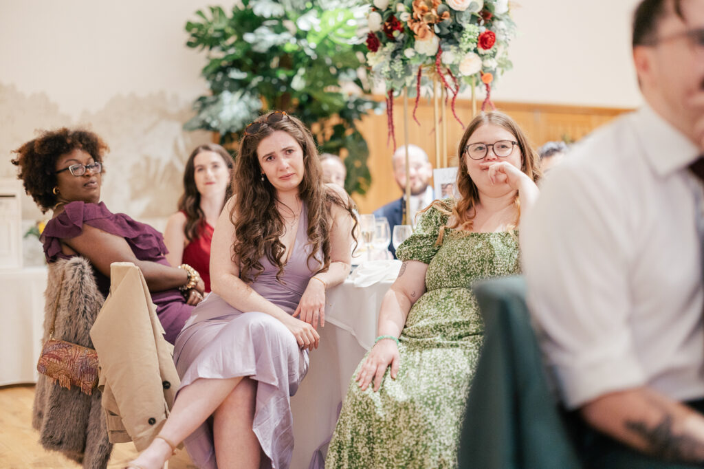 A group of people, mostly women, sit at a table during an indoor event, attentively watching something off-camera. The atmosphere is formal, with floral decorations and elegant attire.