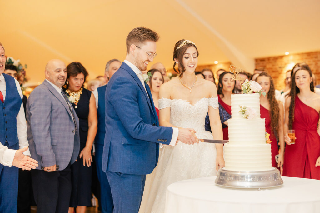 A bride and groom, dressed formally, smile as they cut a tiered white wedding cake together. They are surrounded by well-dressed guests watching and celebrating in a warmly lit room.