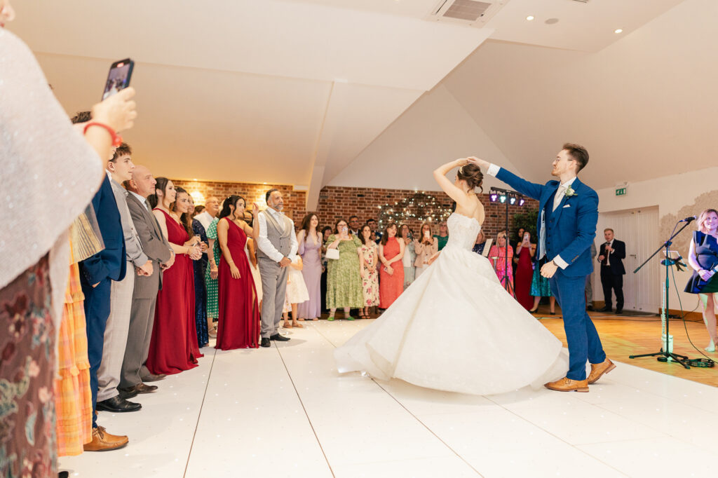 A bride and groom enjoy their first dance in a bright, modern venue surrounded by applauding guests. The bride wears a white gown, the groom a blue suit, and guests watch while someone takes a photo on a phone.