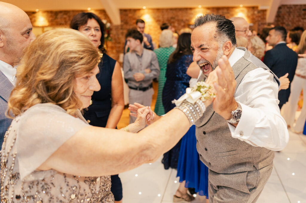 A joyful man in a gray vest laughs and dances energetically with an older woman in a sparkly dress at a lively indoor event, surrounded by other smiling guests.