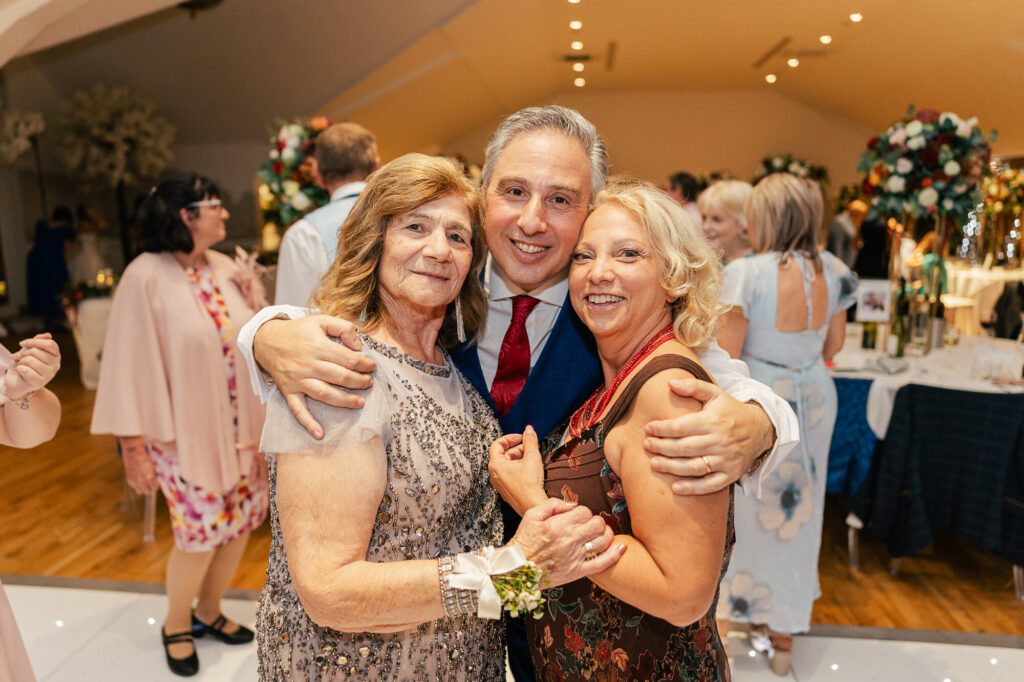 A man in a suit hugs two smiling women at a celebration, likely a wedding reception, with guests and flower arrangements visible in the warmly lit background.