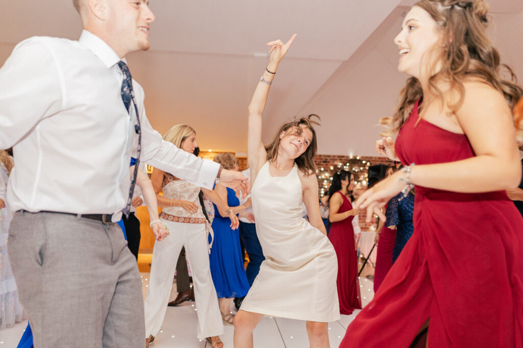Three people are dancing energetically at a wedding or formal event. The woman in the center wears a white dress and raises one arm, while the others, in a shirt and tie and a red dress, smile and enjoy the moment.