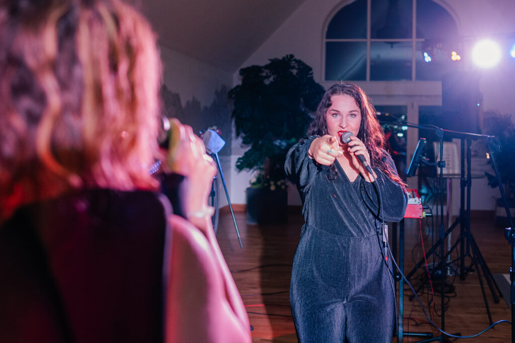 A woman with long hair stands on stage performing with a microphone, pointing toward the camera. Another person with blurred features is in the foreground, also holding a microphone. Stage lights and equipment are visible in the background.