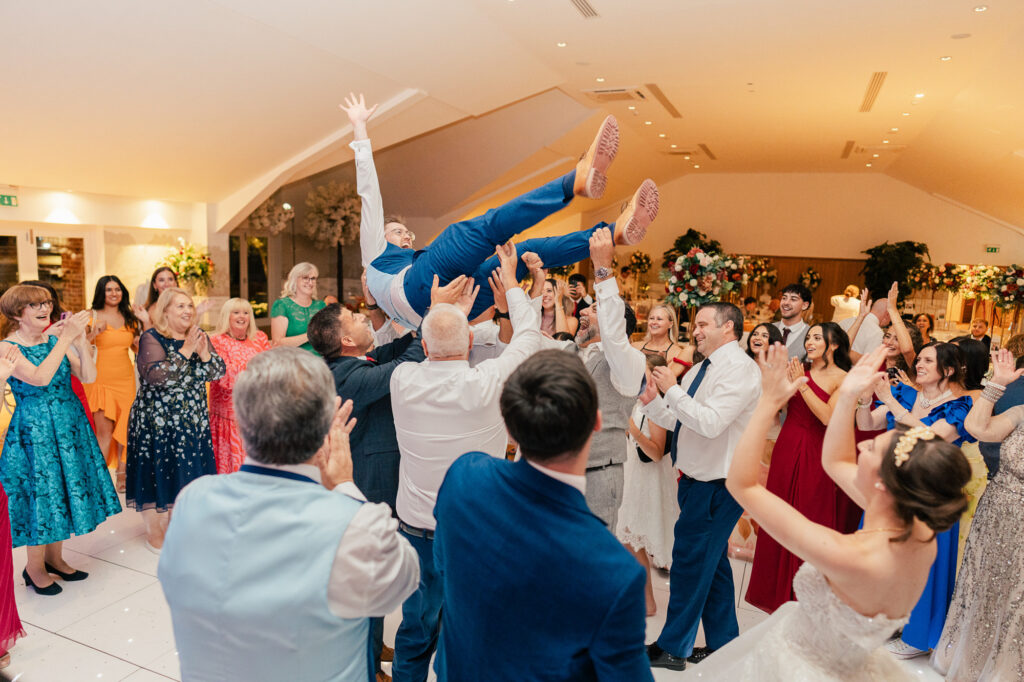 A lively wedding reception where guests joyfully lift a man in a blue suit above the crowd while others cheer and smile, surrounded by colorful dresses and elegant floral decorations.