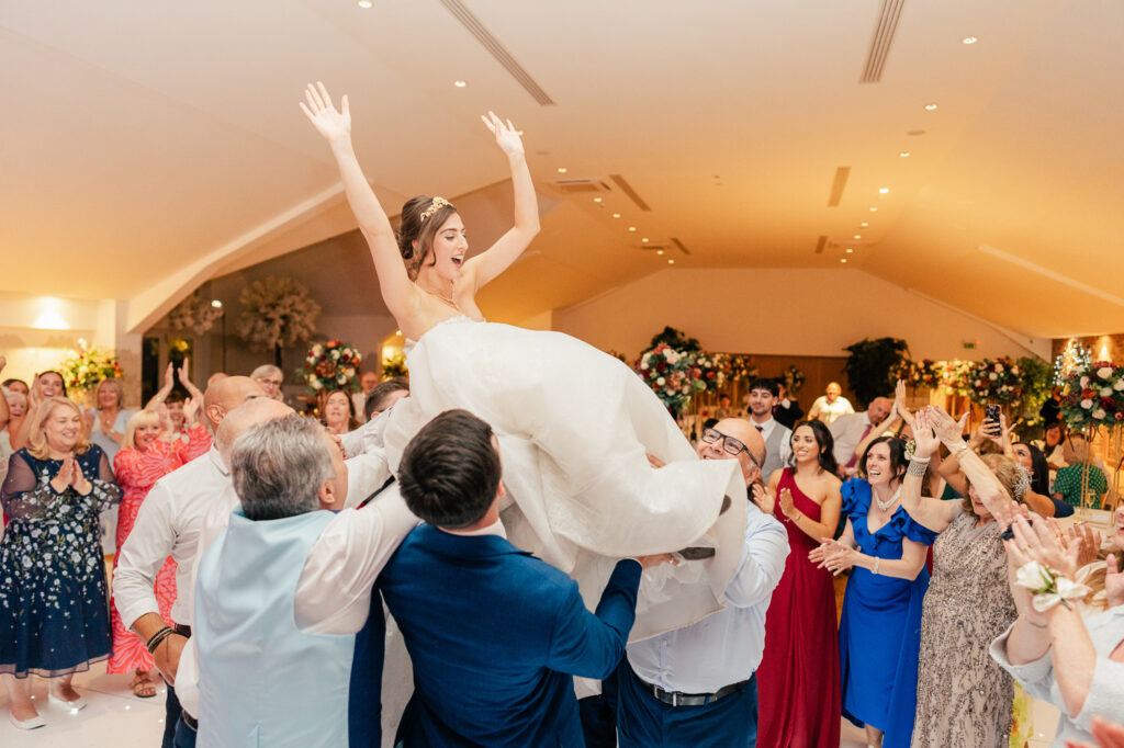 A joyful bride in a white dress is lifted into the air by guests at a lively wedding reception. People around her are smiling, clapping, and celebrating in a brightly decorated hall.