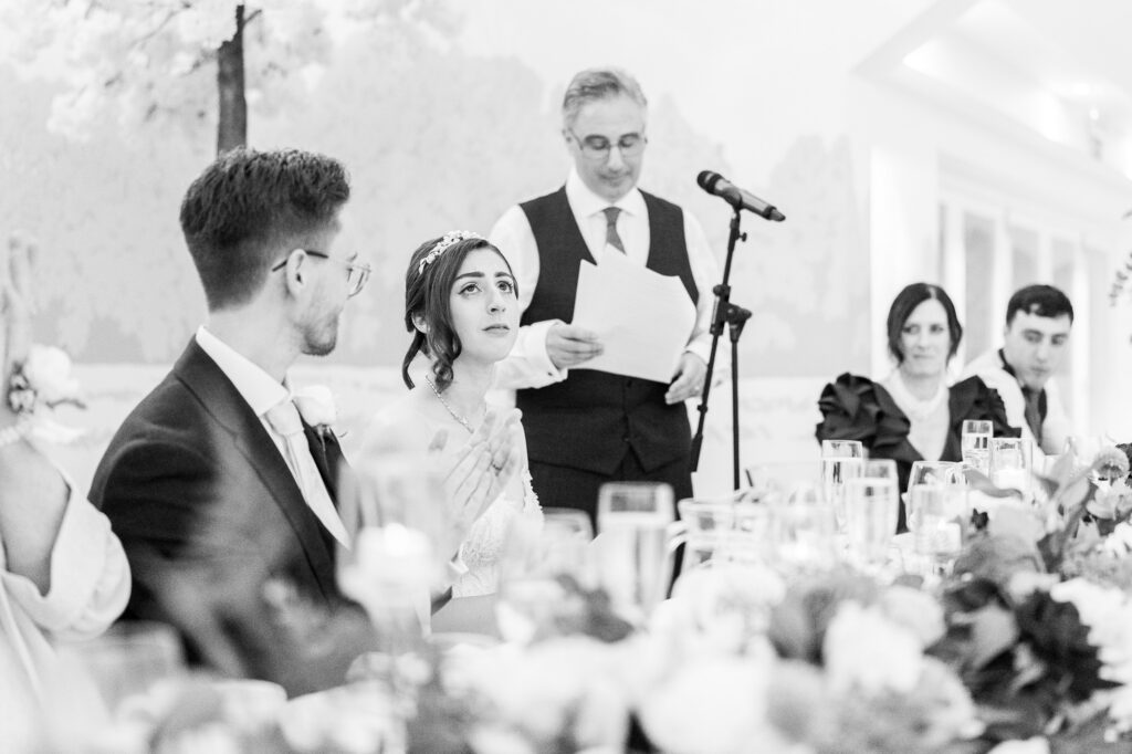 A bride and groom sit at a decorated table during a wedding reception. The bride looks up thoughtfully while a man stands behind them reading from papers into a microphone. Guests are seated around the table.