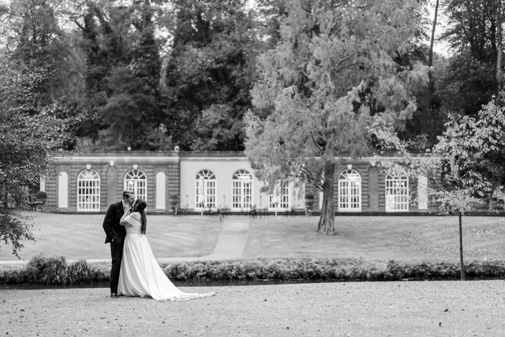 A bride and groom stand closely together in front of a large, elegant building with arched windows, surrounded by trees and a manicured lawn. The photo is in black and white.