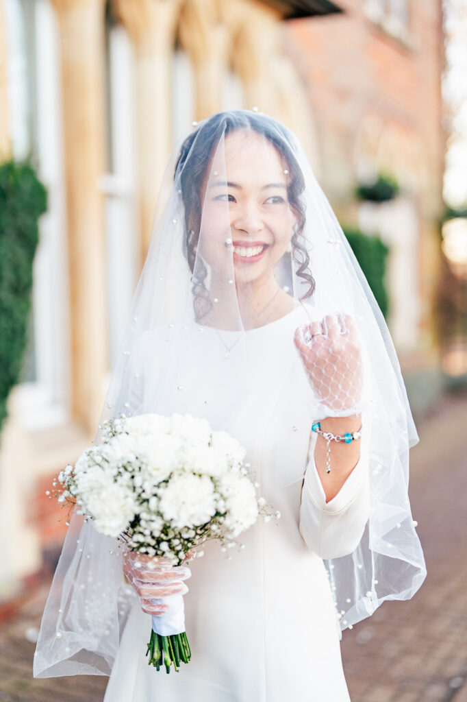 A smiling bride in a white dress and veil holds a bouquet of white flowers, standing outdoors near a stone building with arched windows and greenery in the background.