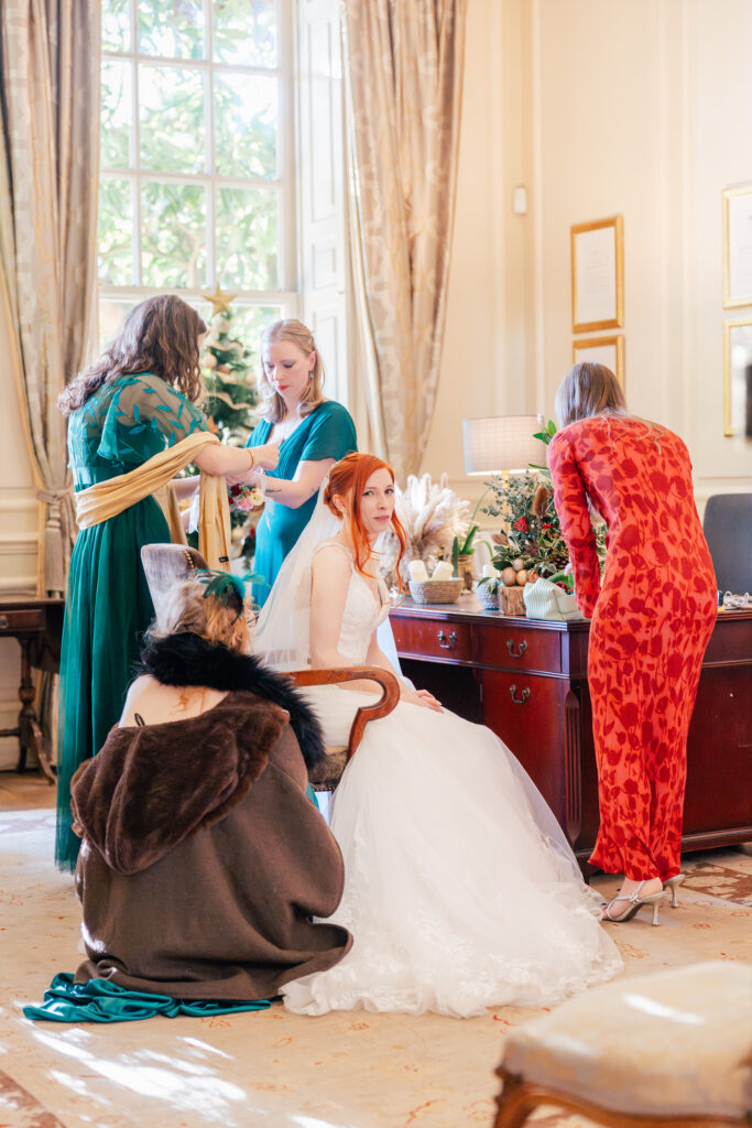 A bride in a white gown sits as three women help adjust her dress and accessories in an elegant, sunlit room; another person in a fur shawl kneels nearby.